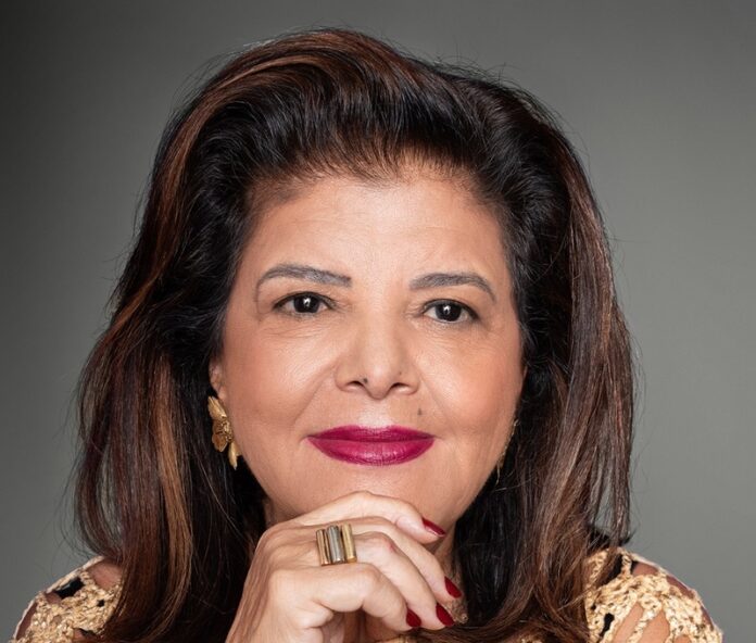 Close-up portrait of a smiling woman with dark hair and red lipstick, resting her chin on clasped hands against a gray studio backdrop.