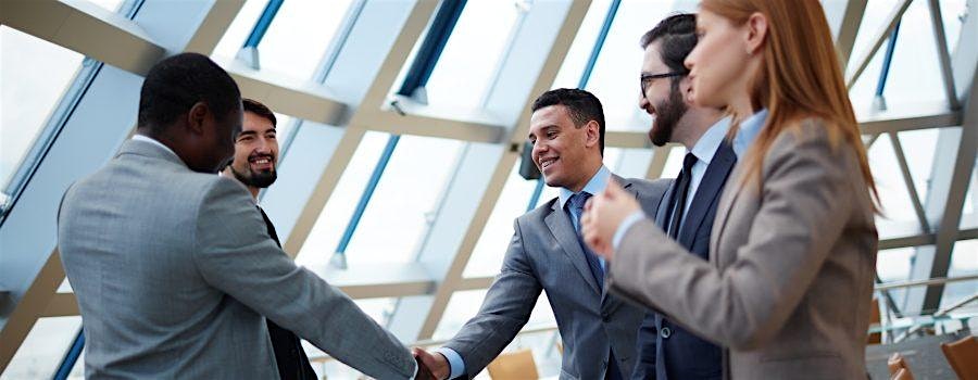 Business professionals in suits shake hands and smile in a bright glass-walled office, signaling a partnership or agreement.