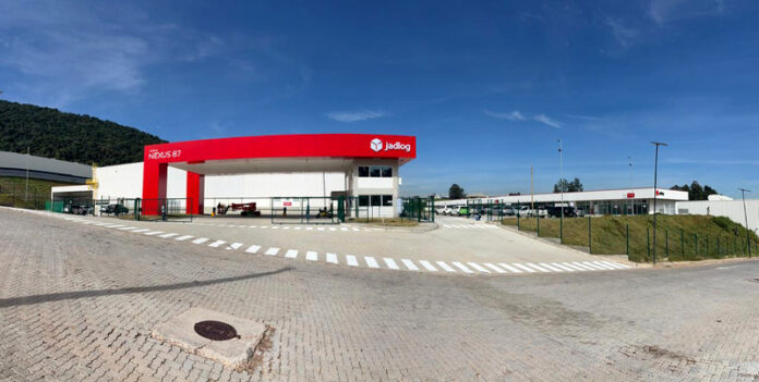 Entrance to a Jadlog logistics center with a red curved canopy, gate, and parking area beyond under a blue sky