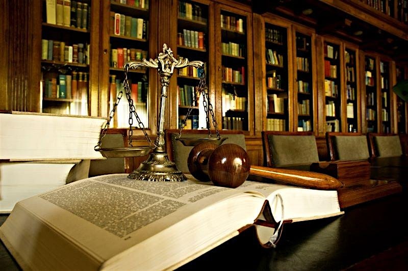 Scales of justice, a wooden gavel, and an open legal tome on a desk in a wood-paneled library.